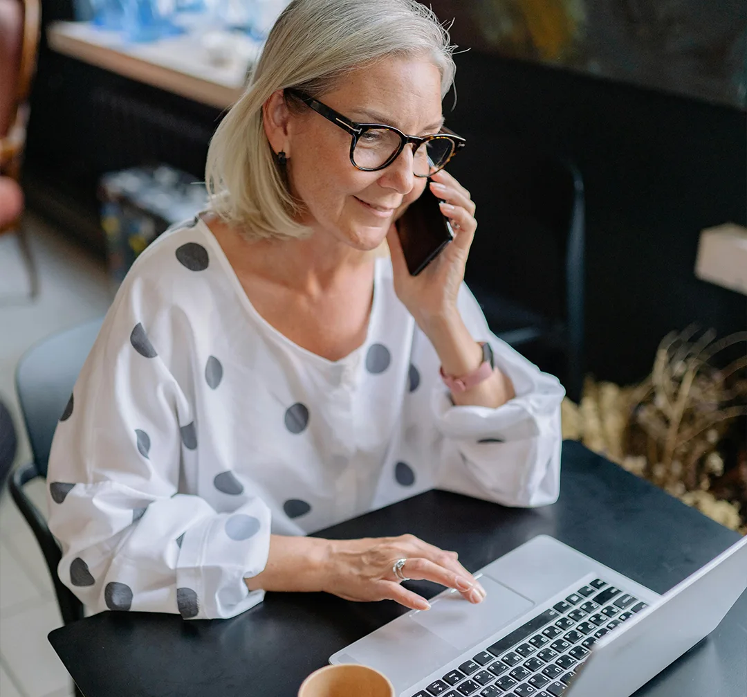 woman in a polka dot blouse in a cafe with a laptop. She holding a phone and smiling
