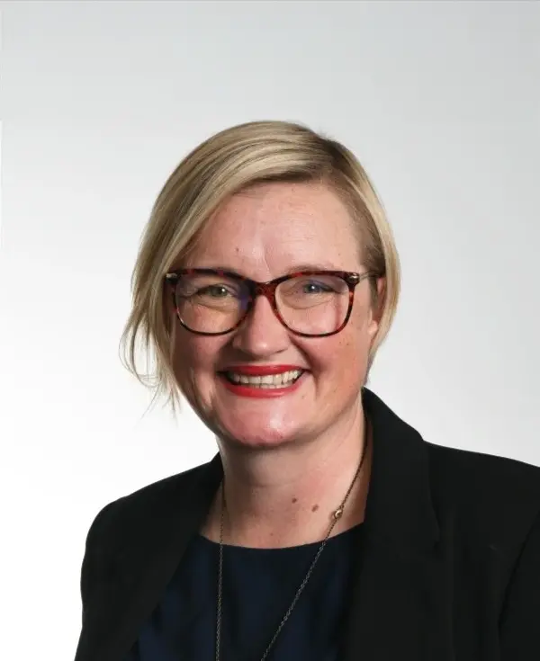 Andrea Nelson in a black suit and wearing glasses. Standing in front of a plain white background