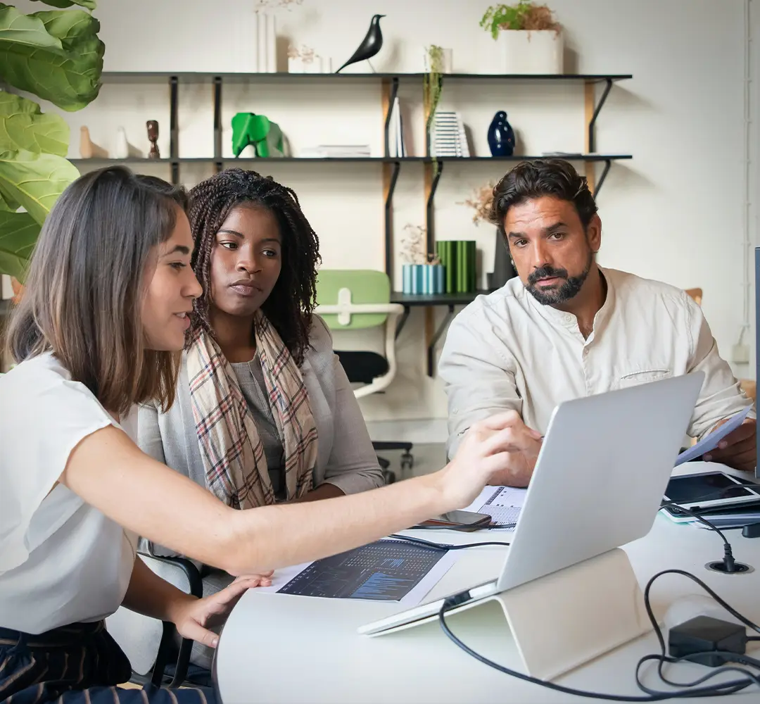 Two women and a man sitting at a table. The women on the left is presenting on a laptop and pointing to her screen
