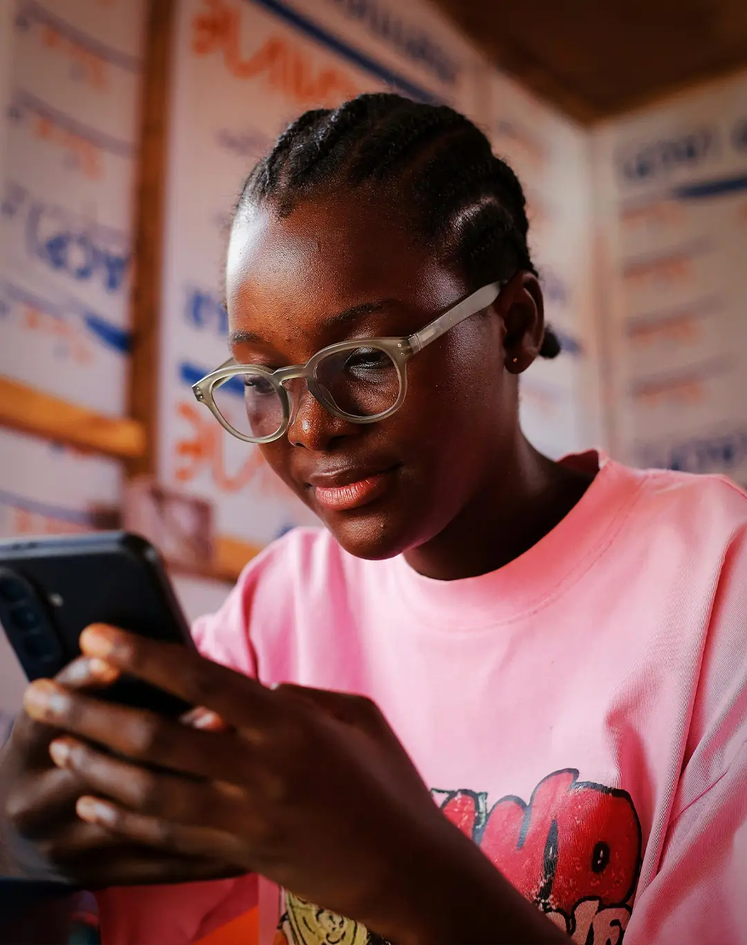 A young Nigerian woman using a smartphone. She's inside and wearing glasses. She's dressed in a pink shirt