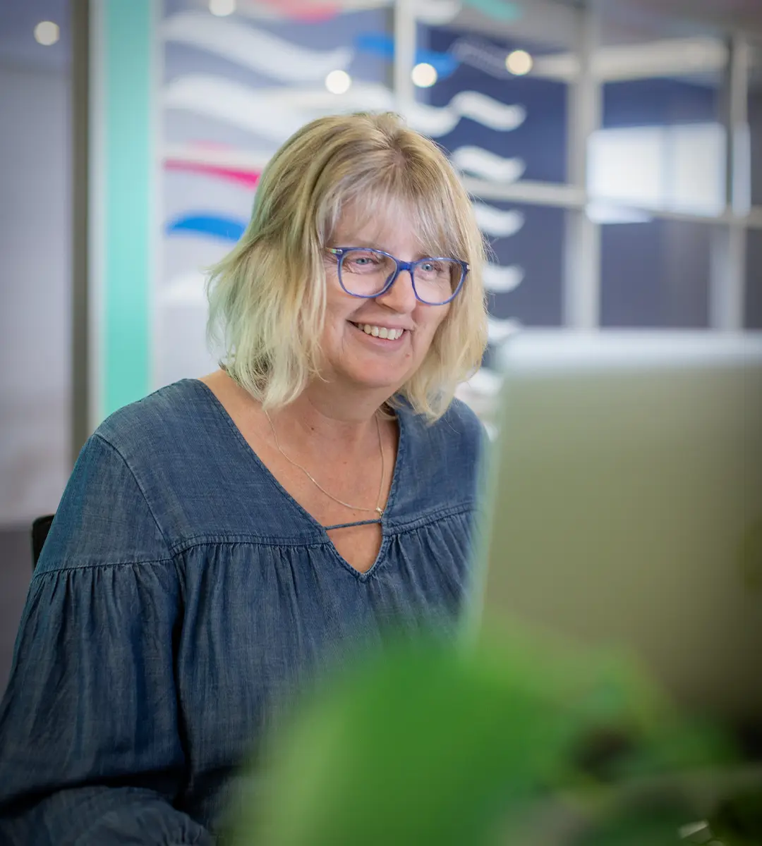 A woman in a denim dress, sitting at a desk. A plant is in the foreground.