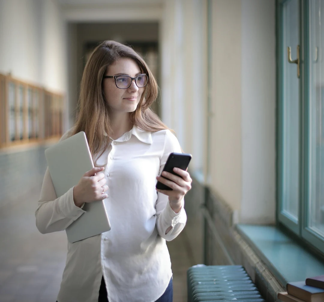 A caucasian woman standing in a hallway wearing a white blouse and glasses. She's holding a phone and laptop and looking out a window