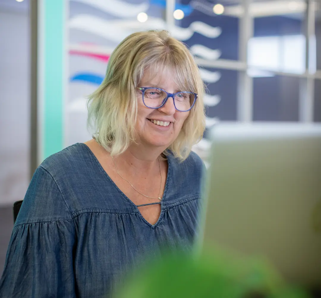 woman in denim dress wearing glasses sitting at a computer and smiling. She's ready to help your nonprofit with bookkeeping