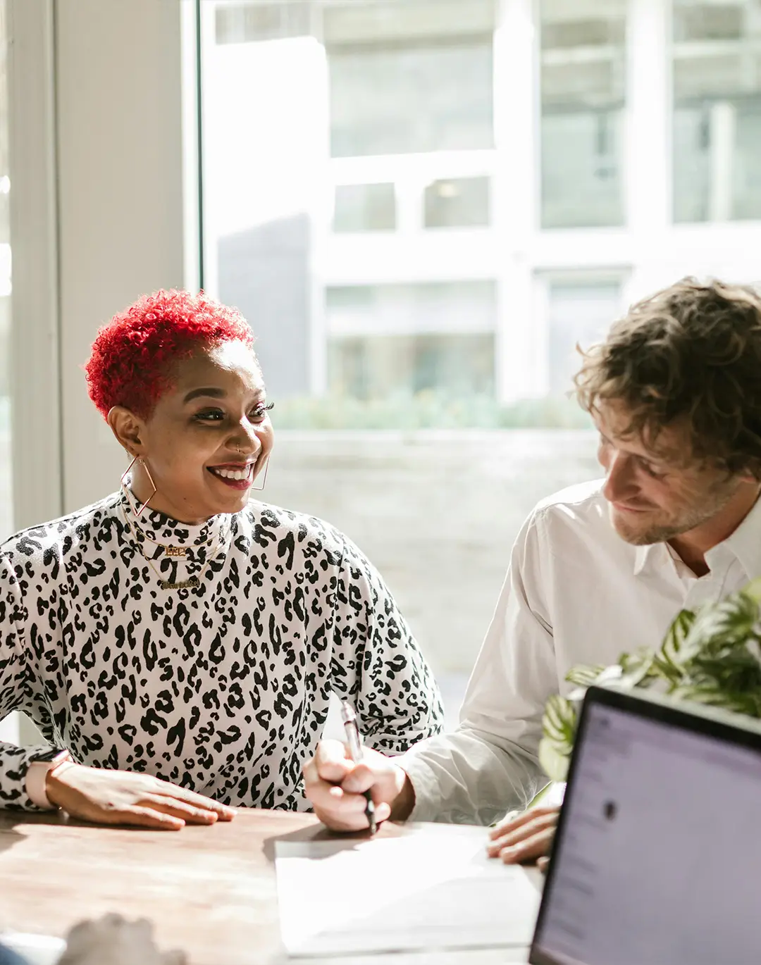 A man and a woman at a conference table, then woman is smiling at the man who is writing on some documents